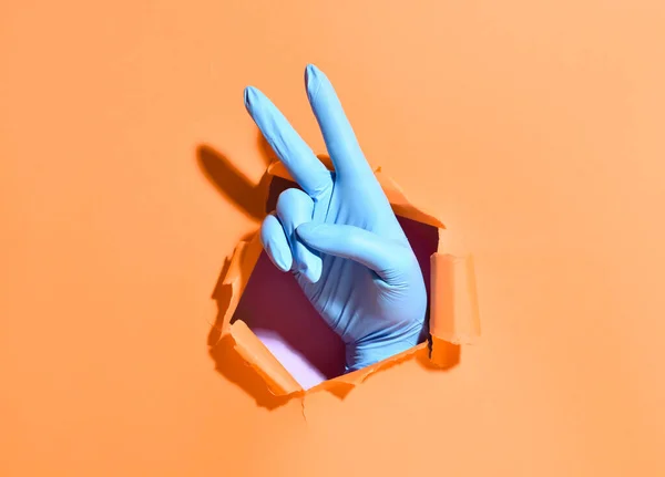 色の背景に紙の穴を通して勝利のジェスチャーを示す医療用手袋で手 — ストック写真