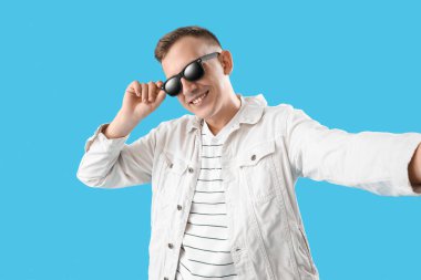 Güneş gözlüklü genç adam mavi arka planda selfie çekiyor.