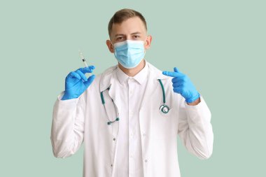 Tıbbi maskeli erkek doktor yeşil arka plandaki şırıngayı işaret ediyor.