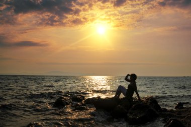 Genç bir kadın denizde kayaların üzerinde duruyor ve bir yerlere bakıyor.