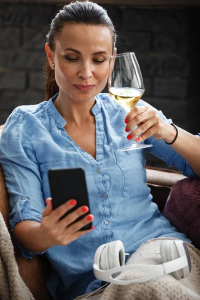 Koncepcja wolnego czasu. Szczęśliwa piękna kobieta rozmawia przez telefon i pije białe wino ze szkła siedząc na kanapie w domu. Kobieta spędza wolny dzień i relaksuje się w domu sama. — Zdjęcie stockowe