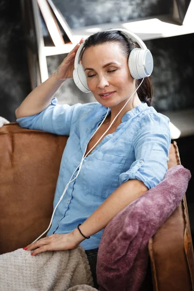 Freizeitkonzept. Glückliche schöne Frau, die die Musik über Kopfhörer hört und auf einer Couch im Haus sitzt. Weibchen verbringen ihren freien Tag und entspannen allein zu Hause. — Stockfoto