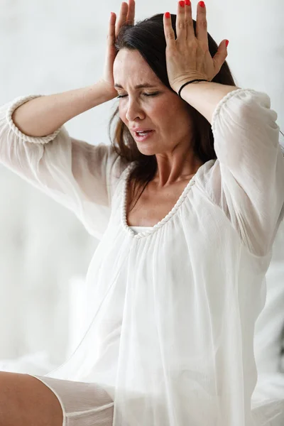 Mujer de mediana edad sentada en una cama blanca en un dormitorio en casa tocándose la cabeza con las manos mientras tiene dolor de cabeza y se siente mal. Sufrimiento de insomnio o migraña —  Fotos de Stock