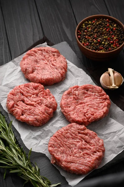 Raw beef burger patties on butcher's wooden board, rosemary and pepper. Black background. View from above.