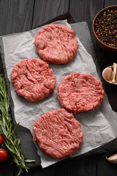 Raw beef burger patties on butcher's wooden board, rosemary and pepper. Black background. View from above.