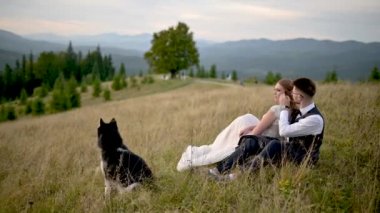Yeni evliler ve köpekleri dağların arka planında çimlerin üzerinde oturuyorlar..