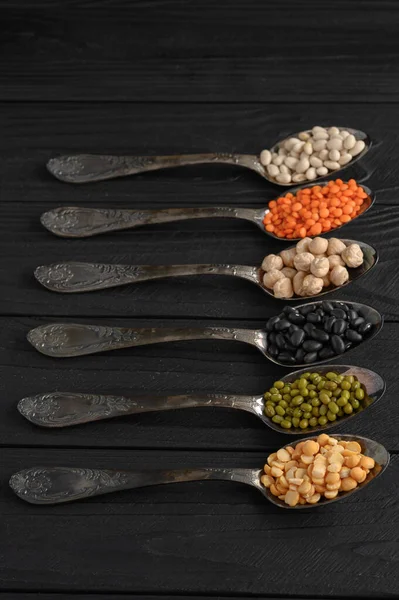 Variety of legumes in old silver spoons on a black wooden background.
