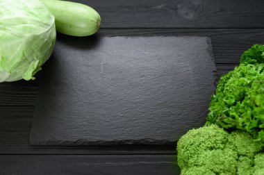 Black stone board on a black wooden background decorated with green vegetables and herbs.