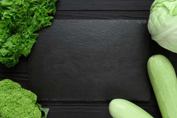 Black stone board on a black wooden background decorated with green vegetables and herbs.