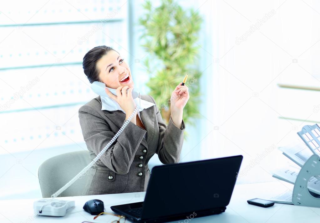 Attractive woman sitting at desk at work on landline phone call, gesturing, smiling.