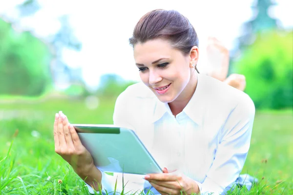 Portrait of a young professional woman laying down on green grass in a city park, using her digital tablet pad with touch screen, smiling. — Stock Photo, Image