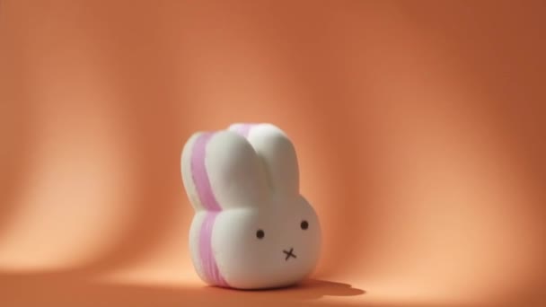 Великий білий і рожевий губчастий кролик падає на помаранчевий фон. Заєць у формі рашистської іграшки відскакує від помаранчевої поверхні в повільному русі. 500 кадрів на секунду — стокове відео