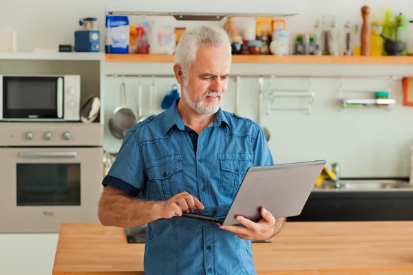 Oude man met laptop zit op de keuken — Stockfoto