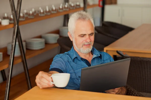 Senior man met laptop zit aan de keuken en drinken koffie — Stockfoto