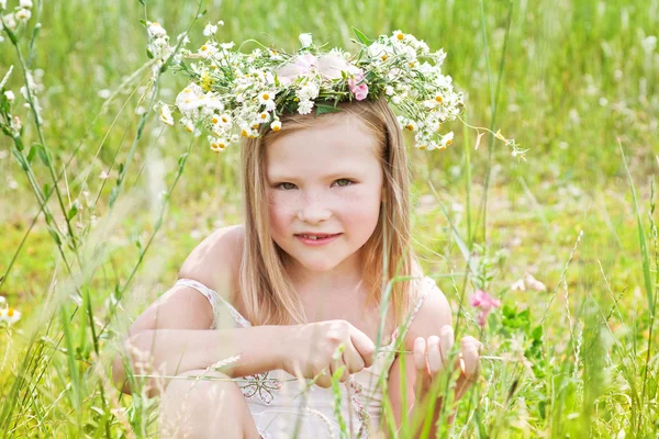 Happy little girl in a wreath of flowers sitting on green grass — Stock Photo, Image