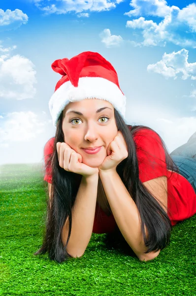 Brunette girl in santa hat is leaning on his elbows on the green — Stock Photo, Image