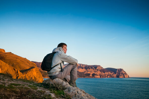 A young man sits on a hill and enjoys sea sunset