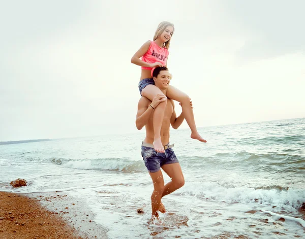 Joven pareja caminando a lo largo de la orilla del mar, chica se sienta en el chico de —  Fotos de Stock