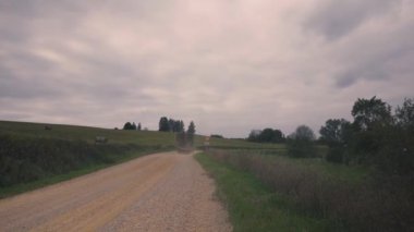 The tractor is carrying hay along a yellow dirt road with gravel. The road turns between hills amid low clouds. Packages of harvested hay lie on the hills. Kurzeme, Latvia
