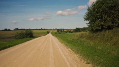 Letonya 'daki tarlalarda sarı çakıl yolu. Yol tepeler arasında bir vadide kıvrılır ve ufkun ötesine geçer. Mavi yaz gökyüzünde beyaz bulutlar var. Orada bir kır evi var.