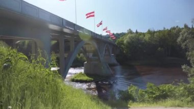 Bridge over the Gauja River in Sigulda on a sunny summer day. The national flags of Latvia are fluttering in the wind on the bridge in the backlight.