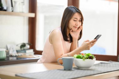 Mutlu gülümseyen Asyalı kadın evde, masada salata kasesiyle akıllı telefon kullanıyor.