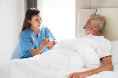 Happy Asian nurse or caregiver holding elderly man patient hand on the bed with care  