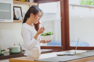 Happy Asian woman eating healthy salad while workinh with laptop on table in kitchen at home
