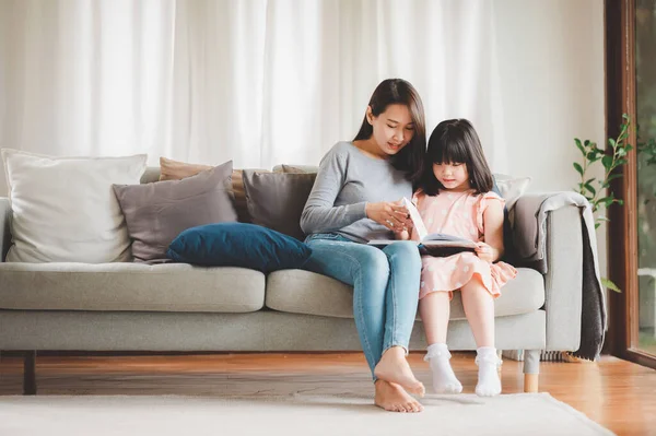 Happy Asian family mother and daughter sitting on sofa reading a book in living room at home