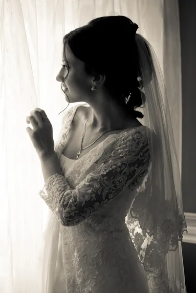 Beautiful young bride standing beside a large window waiting — Stock Photo, Image