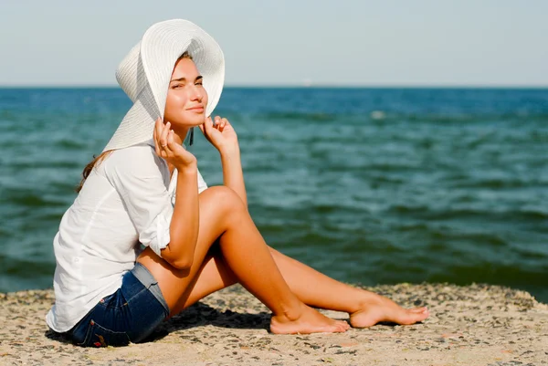 Młoda piękna kobieta siedzi przez błękitne morze i białym kapeluszu — Zdjęcie stockowe