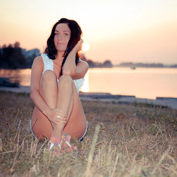 Giovane bella donna seduta sulla riva del fiume al tramonto — Foto Stock