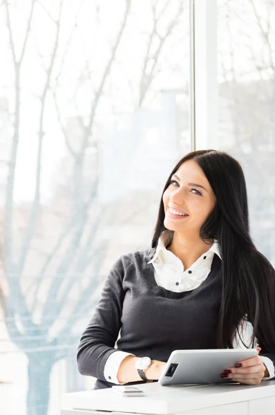 Χαμογελαστή γυναίκα ΝΕΩΝ ΕΠΙΧΕΙΡΗΜΑΤΙΩΝ χρησιμοποιώντας το pc ταμπλετών, ενώ στέκεται χαλαρή κοντά σε παράθυρο στο γραφείο της — Φωτογραφία Αρχείου