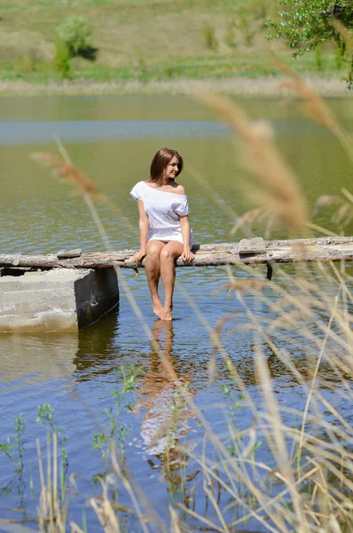 Happy young woman in white dress sitting on pier by river or lake — Stock Photo, Image