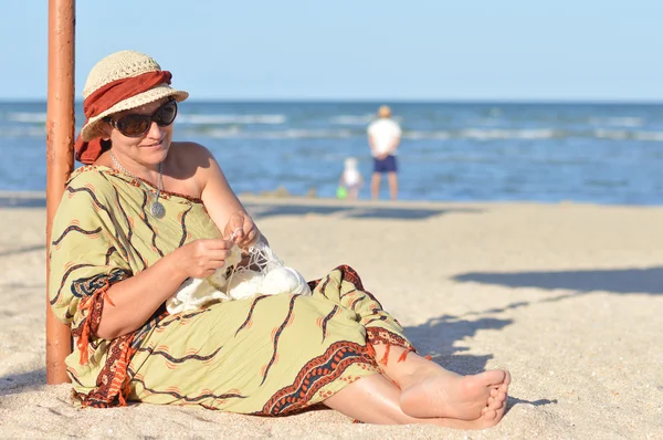 Mujer madura feliz sentada en la playa y tejiendo — Foto de Stock