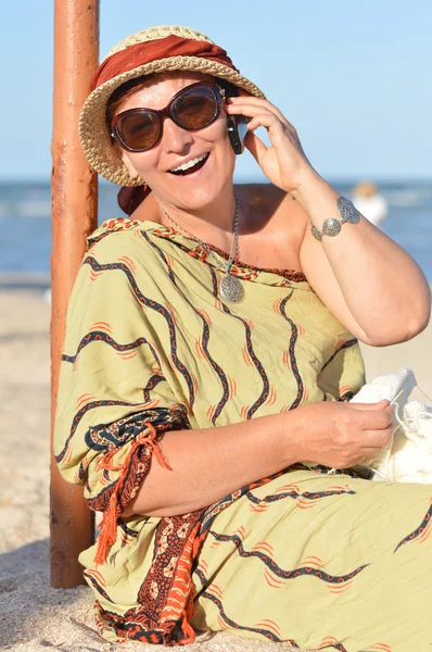 Happy mature woman sitting on beach and talking on mobile phone — Stock Photo, Image