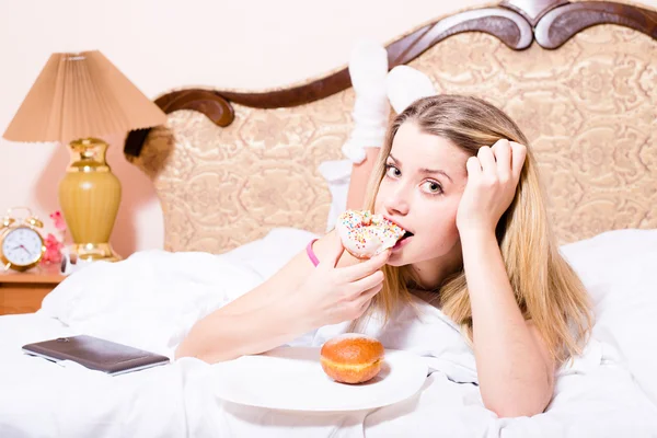 Woman lying in  bed biting color glace donut — Stock Photo, Image