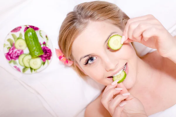 In spa young beautiful attractive girl blond woman lying with slices of cucumber in the hands one piece holding to eye happy smiling & looking at camera — Stock Photo, Image