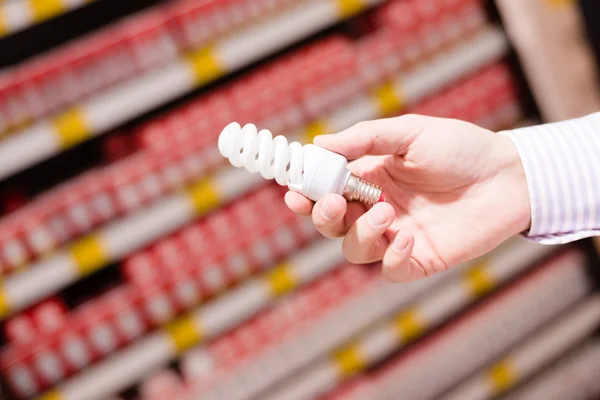 La mano masculina o femenina sostiene o recoge en la tienda iluminación de bajo consumo bombilla ahorrativa primer plano en el fondo del centro comercial DIY — Foto de Stock