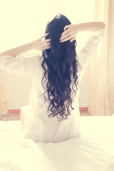 Picture of brunette girl or woman looking in a sunny window holding her head and stretching sitting on white bed — Stock Photo, Image