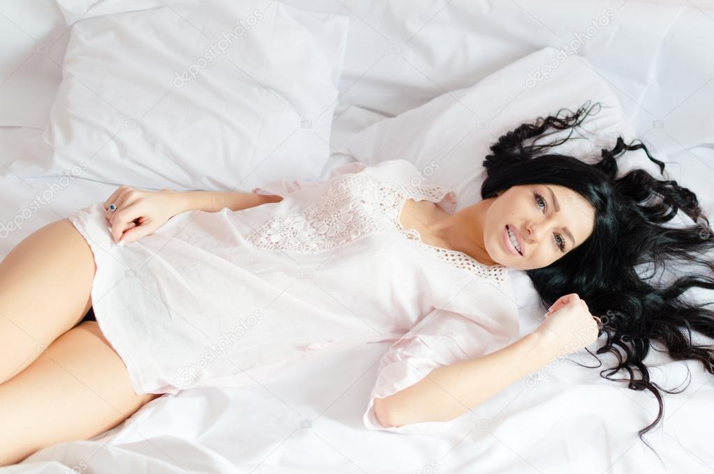 picture of relaxing beautiful young woman brunette girl lying on a white bed in pajamas happy smiling looking at camera portrait