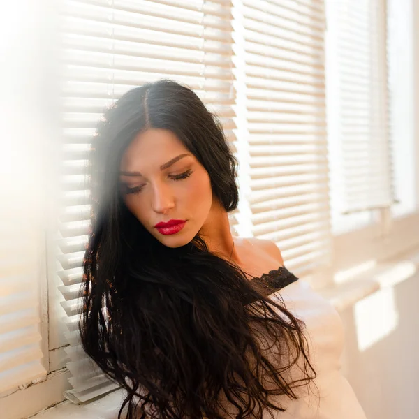 Mooie brunette jonge vrouw met rode lippen op een zonnige verlichting windows achtergrond kijkt neer close-up portret foto — Stockfoto