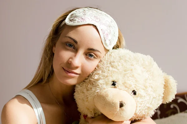 Primo piano ritratto di bella bionda giovane donna con gli occhi blu con benda del sonno sulla testa abbracciando orsacchiotto felice sorridente & guardando l'immagine della fotocamera — Foto Stock
