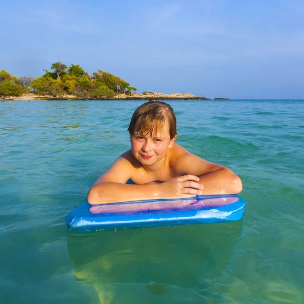 Niño está nadando en su tabla de surf y sonriendo felizmente en un hermoso mar con agua cristalina y cielo azul —  Fotos de Stock