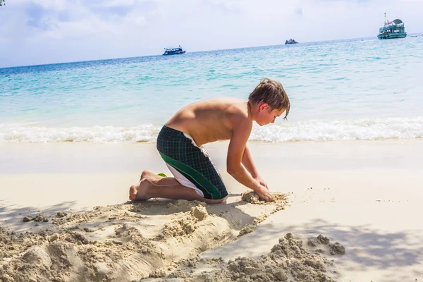 幸せな少年は浜辺で砂から運河を構築します。 — ストック写真