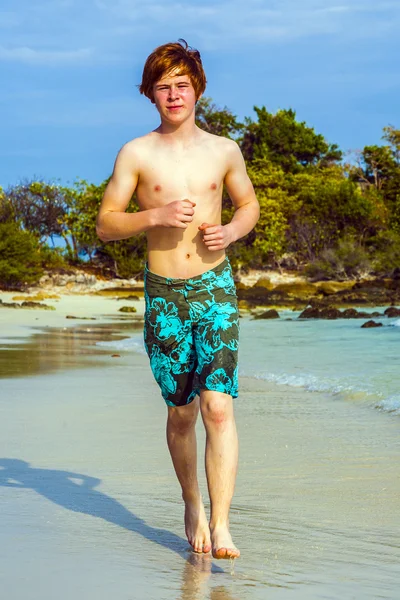 Chico joven con el pelo rojo corriendo a lo largo de la playa tropical — Foto de Stock