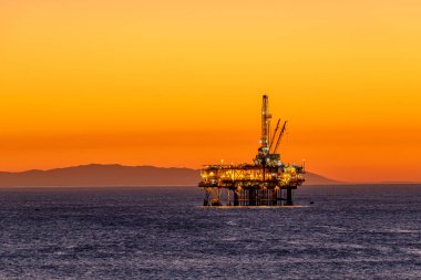 Offshore oil platform off the coast of California against a moody, orange sky as the sun sets behind the rig clipart