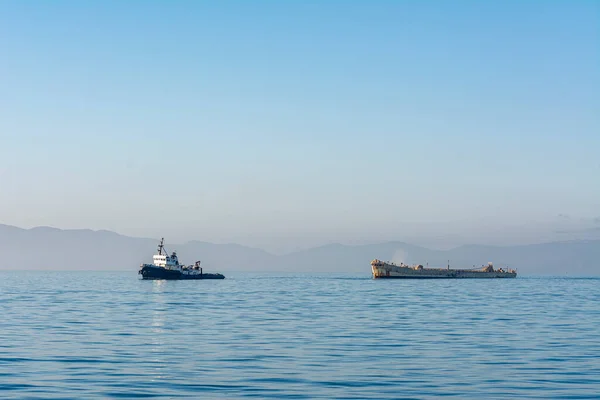 A tugboat tows an old, out of service vessel to a scrap yard through a smoggy, hazy climate