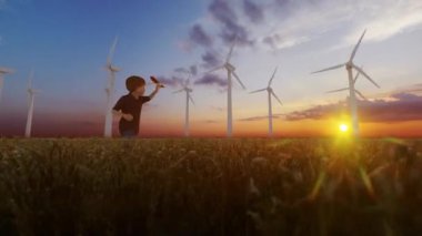 Little boy running in a wheat field with wind turbines on the background at sunrise, 4K