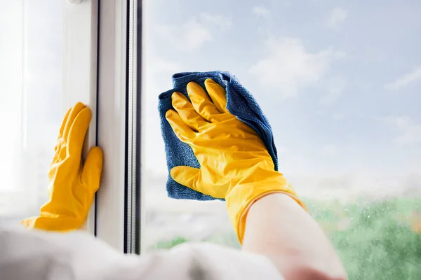 Man in yellow rubber gloves cleaning window glasse with cleaner spray detergent and squeegee or rag at home or office, copy space. Housework and housekeeping, home hygiene. Blue sky view, plastic window frames